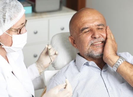 Older man in dental chair holding his cheek in pain