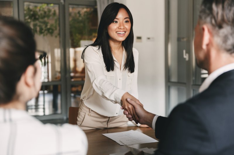 Woman smiling with teeth shaking hands with man in suit across table from her in office setting